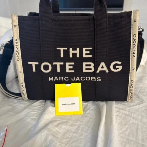 Svart The Tote Bag från Marc Jacobs - Säljer en svart The Tote Bag från Marc Jacobs i canvas med vita detaljer och bred axelrem. Väskan har tydlig logga framtill och kontrasterande kanter med text. Perfekt storlek för att få plats med allt du behöver och riktigt snygg att bära crossbody eller i handen.