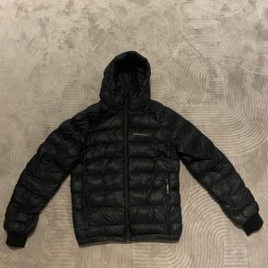 Svart dunjacka Peak Performance S - Svart dunjacka från Peak Performance med huva och dragkedja framtill. Den är i storlek S och är perfekt om man vill ha en varm men också snygg jacka till vintern. Finns 2 skador på den men båda är fixade hos skräddare. 1 av de är ett litet hål på armen(bild 4) medan den andra sitter på luvan men är ej synligt! Nypris är 3199kr