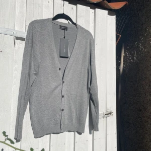 Grå kofta/cardigan från Zara - Snygg grå kofta från Zara med v-ringad hals och knappar framtill. Långärmad och tillverkad i mjuk bomull, perfekt för lager-på-lager. Enkel och stilren design som passar till jeans eller chinos.