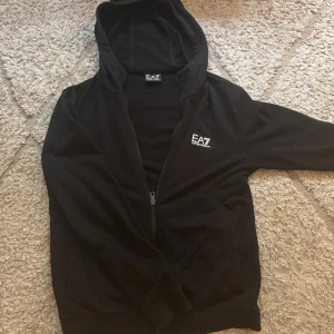 Svart EA7 hoodie med dragkedja - Svart hoodie från EA7 Emporio Armani med vit logga på bröstet och huva. Tröjan har dragkedja framtill, långa ärmar och ribbade muddar. Perfekt för en chill och sportig look. Materialet är mjukt och känns skönt mot huden.