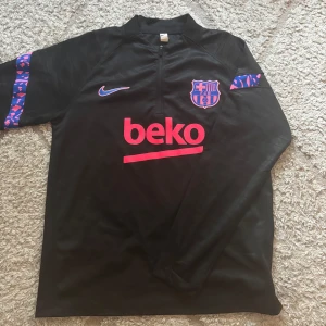 Svart långärmad FC Barcelona träningströja - Svart långärmad FC Barcelona träningströja från Nike med halv dragkedja. Tryckt klubbmärke och Nike-logga i lila och rosa, samt sponsortryck i rosa. Ärmarna har blå och rosa leopardmönstrade detaljer. Tillverkad i lätt funktionsmaterial.