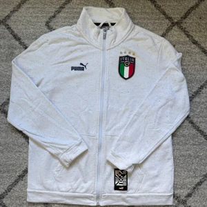 Vit Puma Italia ziptröja M - Säljer en vit långärmad ziptröja från Puma med Italia-emblem och coola färgprickar. Tröjan har hel dragkedja, ståkrage och tryck både fram och bak. Perfekt för dig som gillar fotboll och vill ha en sportig look.