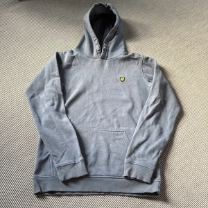 Lyle & Scott - Grå hoodie från Lyle & Scott Junior - Grå hoodie från Lyle & Scott Junior med klassisk gul logga på bröstet. Tröjan har huva, känguruficka och ribbade muddar vid ärmslut och nederkant. Perfekt för en chill och avslappnad stil.