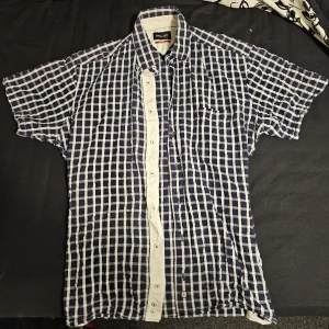 Blårutig kortärmad skjorta L - Kortärmad skjorta L i blått och vitt rutigt mönster. Skjortan har klassisk krage, knappstängning framtill och en bröstficka. Perfekt för en avslappnad och cool stil under varmare dagar.