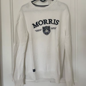 Vit stickad tröja från Morris - Snygg vit stickad tröja från Morris med broderad logga och emblem på bröstet. Tröjan har rund halsringning, ribbade muddar och lång ärm. Perfekt för dig som gillar klassisk och stilren design.