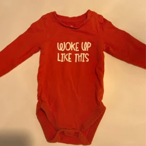 Röd långärmad body med text - Röd långärmad body i mjuk bomull med vit text på bröstet: 'WOKE UP LIKE THIS'. Praktiska knappar i grenen och vid axeln för enkel påklädning. Perfekt för en chill och skön stil.