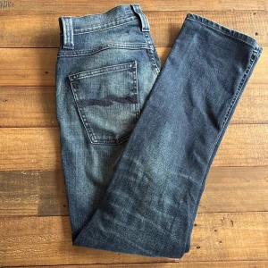 Nudie Jeans blå raka jeans - Säljer ett par klassiska blå jeans från Nudie Jeans med raka ben och snygga slitningar. Jeansen har fem fickor, tydliga sömmar och Nudie-logga på bakfickan. Tillverkade i bomull med en cool tvättad look och normal passform.