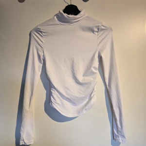 Vit polotopp med rynkade sidor - Säljer en stilren vit polotopp med långa ärmar och rynkade detaljer på sidorna. Toppen har en hög krage och är figurnära i modellen, perfekt att styla med jeans eller kjol. Materialet känns mjukt och stretchigt.