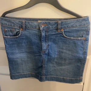 Blå jeanskjol med klassisk passform - Snygg blå jeanskjol med klassisk femficksmodell och knapp- samt dragkedjestängning framtill. Kjolen har en rak, kort siluett och tydliga sömmar i kontrastfärg. Perfekt för dig som gillar en enkel och tidlös denimlook.