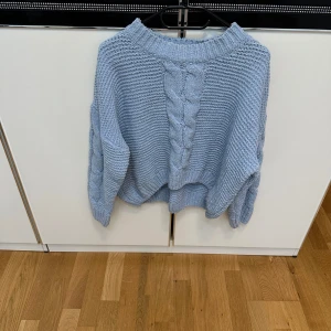 Ljusblå stickad tröja med flätmönster - Supermysig ljusblå stickad tröja med grovt flätmönster både fram och på ärmarna. Tröjan har en bred ribbad halsringning och är lite oversized i modellen.