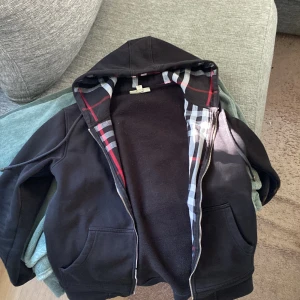 Svart burberry hoodie  - Svart hoodie med hel dragkedja och snörning i huvan. Mjukt bomullsmaterial och klassisk passform. Fodret har rutigt mönster i svart, vitt och rött. Han på bilden ä 182