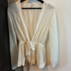 Vit omlottblus med knytband från NAKD - Superfin vit omlottblus från NAKD med plisserat tyg och långa ärmar. Blusen har ett brett satinband att knyta i midjan och v-ringad halsringning. Perfekt för dig som gillar romantiska och luftiga plagg.