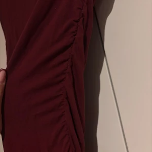 Vinröd långklänning med rynkade sidor - Säljer en vinröd långklänning i mjukt tyg med rynkade detaljer längs sidorna som framhäver kroppens form. Klänningen har en enkel, elegant design med boatneck halsringning och är ärmlös. Perfekt för dig som gillar stilrena och bekväma plagg med en twist.❤️
