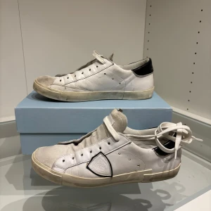 Philippe Model vita sneakers med svart detalj - Snygga vita sneakers från Philippe Model med svart hälkappa och klassisk logga på sidan. Skorna har rund tå, snörning och platt sula. Tillverkade i skinn och mocka med kontrastdetaljer och en vintage-inspirerad look. Perfekta för dig som gillar stilrena men ändå unika sneakers.