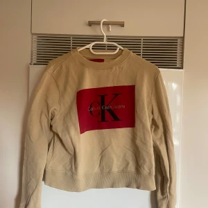 Beige sweatshirt från Calvin Klein Jeans - Säljer en beige sweatshirt från Calvin Klein Jeans med stort rött logotryck framtill. Tröjan har rund halsringning, långa ärmar och ribbade muddar vid ärmslut och nederkant. Perfekt för en chill och stilren look.