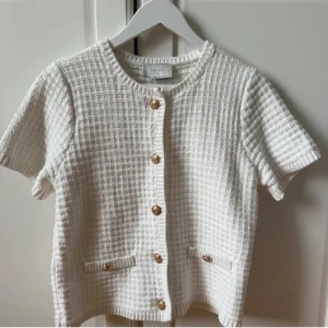 Vit kofta med guldknappar från Neo Noir - Kortärmad vit kofta från Neo Noir i stickad struktur med snygga guldknappar framtill och två små fickor. Perfekt för lager-på-lager och ger en chic vibe till din outfit.✨🤍💛