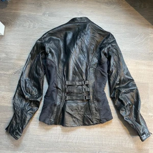 Svart skinnjacka med spännen - Snygg svart skinnjacka/motorcykeljacka från joerocket med coola spänndetaljer i ryggen och dragkedjor vid ärmslut och fickor. Jackan har figurnära passform, bra skick men tecken på användning