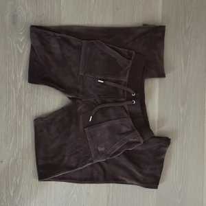 Brunt Juicy Couture sett  - Mysiga bruna mjukisbyxor och hoodie från Juicy Couture i mjuk velour. Byxorna och tröjan har en dragsko, fickor samt en bred mudd i midjan. Perfekt om man vill klä sig Bekvämt och mysigt. 
