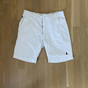 Vita Polo Ralph Lauren shorts - Snygga vita shorts från Polo Ralph Lauren med klassisk passform och broderad logga på benet. Tillverkade i mjuk bomull med fickor fram och bak samt bälteshällor. Perfekta för en fräsch och stilren sommarlook.30 midja men väldigt tajt om du ska ha häng och har den storleken. Passar 175-180. 