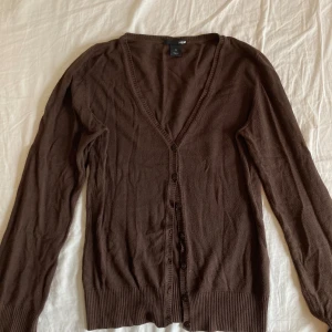 Brun kofta från H&M med knappar - Mysig brun kofta från H&M med v-ringning och knäppning framtill. Långärmad modell med ribbade muddar och nederkant. 
