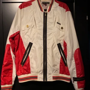 Wind brakerDolce & Gabbana - Snygg bomberjacka från Dolce & Gabbana i vitt och rött med svarta detaljer. Jackan har dragkedja framtill, två fickor med dragkedja och ribbade muddar med röd och svart rand. Materialet är glansigt och lätt, perfekt för en sportig look.