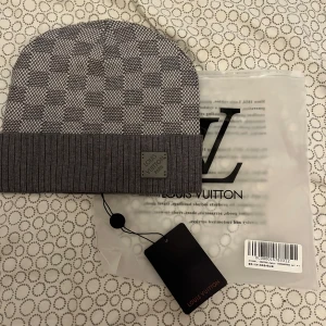 Grå rutig mössa från Louis Vuitton - Exklusiv grå mössa från Louis Vuitton med klassiskt rutigt mönster och ribbad uppvikt kant. Mössan har en diskret Louis Vuitton-patch framtill och är tillverkad i mjukt material som känns lyxigt mot huden. Perfekt accessoar för att lyfta din vinterstil.