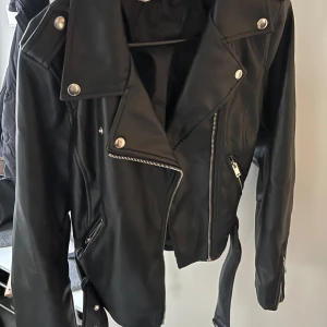 Svart skinnjacka med dragkedjor - Säljer en svart skinnjacka i klassisk bikerstil med silvriga dragkedjor, knappar och bälte i midjan. Jackan har långa ärmar och coola detaljer som bred krage och fickor med dragkedja. Nypris 799kr.
