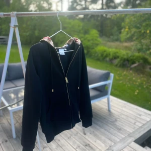 Svart hoodie från Burberry - Svart hoodie från Burberry med klassiskt rutigt foder i huvan. Tröjan har dragkedja framtill, snörning i huvan och ribbade muddar. Perfekt för dig som gillar stilrena plagg med en twist av lyx. Ikoniskt Burberry-mönster i beige, rött och svart på insidan av huvan.