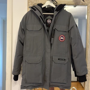 Grå Canada Goose Expedition dunjacka - Säljer en grå Canada Goose Expedition dunjacka med stor huva, flera praktiska fickor och klassisk logga på bröstet. Jackan har svarta muddar vid ärmsluten och är perfekt för kalla vinterdagar. Väldigt fint skick. Nypris 11000 kr 