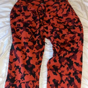 Nike camo mjukisbyxor röd/svart M - Nike mjukisbyxor i röd och svart camo-mönster med svarta snören i midjan. Byxorna har en loose passform och är tillverkade i mjukt bomullsmaterial. Perfekta för chill eller streetwear, med Nike-logga på låret.