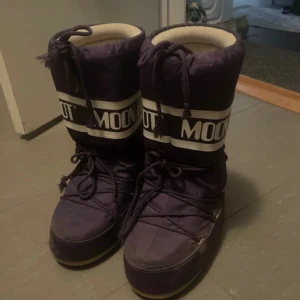 Lila Moon Boot vinterstövlar - Moon Boot vinterstövlar i mörklila med vit insida och bred vit logotypdetalj runt skaftet. Stövlarna har snörning framtill och är tillverkade i syntetmaterial, perfekta för kalla vinterdagar. Klassisk rund tå och platt sula för skön komfort. Skriv vid mer frågor 