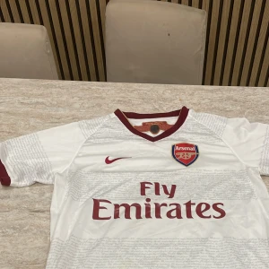 Vit Arsenal fotbollströja Nike S - Snygg vit Arsenal fotbollströja från Nike med vinröda detaljer och klubbmärke på bröstet. Tröjan har korta ärmar, V-ringad krage och diskret textmönster över hela tyget. Materialet är lätt och andas, perfekt för match eller träning.