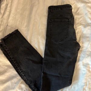 Svarta jeans med rå kant från Gina Tricot - Svarta jeans från Gina Tricot med hög midja och rå, fransig kant vid benslutet. Klassisk femficksmodell i stretchig denim som sitter snyggt och bekvämt. Perfekta för dig som gillar en enkel men trendig look.