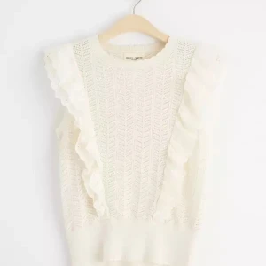 Vit spetsig blus - Supersöt vit blus från Holly & Whyte med broderade volangdetaljer längs axlarna och ett stickat, hålmönstrat tyg. Ärmlös modell med ribbad midja och romantisk känsla. Perfekt för dig som gillar en feminin och trendig stil.