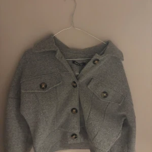 Grå overshirt med knappar - Säljer en grå overshirt från Bik Bok med stora bruna knappar och två bröstfickor. Jackan är croppad och har en boxig passform med klassisk krage. Perfekt att slänga över en hoodie eller t-shirt för en chill look.