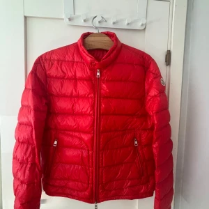 Röd dunjacka från Moncler - Säljer en röd dunjacka från Moncler med klassisk quiltad design och hög krage. Jackan har dragkedja framtill, två fickor med dragkedja och Moncler-logga på ärmen. Perfekt för kyliga dagar och ger en riktigt clean look.