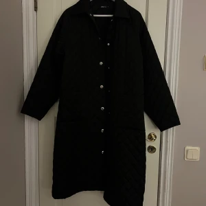 Svart quiltad lång jacka från Gina Tricot - Säljer en svart lång quiltad jacka från Gina Tricot. Jackan har knappar framtill, krage och stora fickor. Perfekt för höst  med sin stilrena look och bekväma passform.