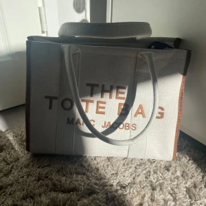 Marc Jacobs The Tote Bag beige - Snygg och rymlig shoppingväska från Marc Jacobs i beige canvas med bruna detaljer och tydlig logga framtill. Väskan har både handtag och en avtagbar axelrem med svartvita ränder. Praktisk dragkedja upptill och svart läderpatch på baksidan. Inte äkta!