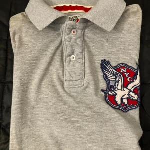 Grå pikétröja Hilfiger Denim NYC - Snygg grå pikétröja från Hilfiger Denim med klassisk krage och knäppning framtill. Stor NYC-patch med örn i rött, vitt och blått på bröstet. Röd och vit detalj på insidan av kragen. Perfekt för dig som gillar sportig och stilren look.