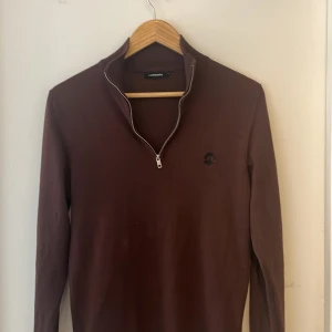 Vinröd half zip tröja från J.Lindeberg - Säljer en vinröd långärmad tröja från J.Lindeberg med half zip-dragkedja och diskret svart logga på bröstet. Tröjan är i mjukt material och har en klassisk, stilren look som passar perfekt till lager-på-lager under höst och vinter.