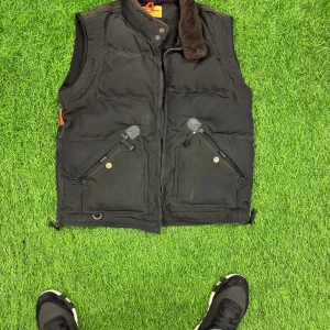 Svart dunväst från Parajumpers XLL - Svart dunväst från Parajumpers i storlek XL med bruna detaljer på kragen. Västen har två snedställda fickor med dragkedja och knapp, samt meshfickor på insidan. Stängs med dragkedja och tryckknapp vid halsen. Perfekt för lager-på-lager under kyliga dagar.