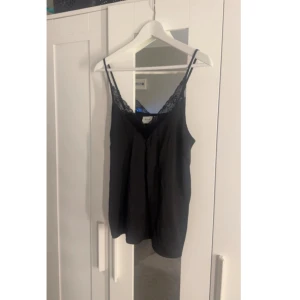 Svart linnetopp med spetsdetalj - Snygg svart linnetopp med tunna axelband och v-ringning dekorerad med spets. Toppen har en lös passform och är perfekt för att skapa en stilren look. Enkel att matcha med både jeans och kjol.