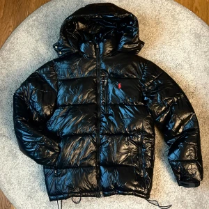 Svart pufferjacka Polo Ralph Lauren - Snygg svart pufferjacka från Polo Ralph Lauren med glansig finish, röd broderad logga på bröstet och avtagbar huva. Jackan har dragkedja framtill, flera fickor med dragkedja och justerbara muddar. Perfekt för kalla dagar, ett av spännbanden som går till höger ficka har gått sönder, kan skicka bild
