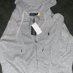 Grå hoodie från Polo Ralph Lauren - Snygg ljusgrå hoodie från Polo Ralph Lauren, helt ny och oanvänd med dragkedja och klassisk logga broderad på bröstet. Tröjan har huva med snörning och ribbade muddar. Perfekt för en chill och stilren look.