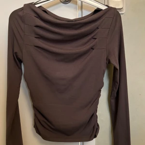 Ny Brun långärmad topp Gina Tricot - Säljer en brun långärmad topp från Gina Tricot med snygga draperade detaljer vid halsringningen. Toppen har en figurnära passform och är tillverkad i mjukt bomullsmaterial. Perfekt att styla med jeans eller kjol för en trendig look.