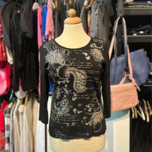 Svart topp med paisleymönster GR Design - Svart långärmad topp från GR Design i 100% bomull. Framsidan har ett detaljerat paisley- och blomliknande mönster i vitt och grått samt tryckt text. Toppen har rund halsringning och en figurnära passform. 