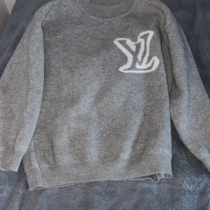 Grå stickad Louis Vuitton tröja - Snygg grå stickad tröja från Louis Vuitton med stor vit LV-logga både fram och bak. Tröjan har rund halsringning, ribbade muddar och är långärmad. Perfekt för dig som vill ha en stilren och exklusiv look.