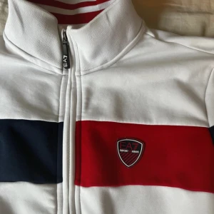 Vit EA7 track jacket med röd och marinblå detalj - Snygg vit track jacket från EA7 Emporio Armani med breda ränder i rött och marinblått över bröstet. Jackan har hög krage med röda ränder på insidan, dragkedja framtill och EA7-logga på bröstet. Perfekt för en sportig och clean look.