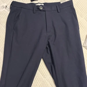 Mörkblå kostymbyxor Jack & Jones - Snygga mörkblå kostymbyxor från Jack & Jones med klassisk rak passform och midwaist. Byxorna har bälteshällor, knapp och dragkedja framtill samt sidfickor. Materialet är mjukt och stretchigt för extra komfort.
