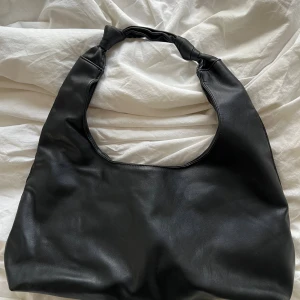 Svart axelväska i skinn imitation med knutdetalj - Stilren svart axelväska i skinnimitation med unik knutdetalj på axelbandet. Väskan har ett rymligt huvudfack med dragkedja och en liten ficka inuti. Perfekt för dig som gillar minimalistisk och trendig look. Har en liten slitning på axelbandet, vänligen se bild 3.